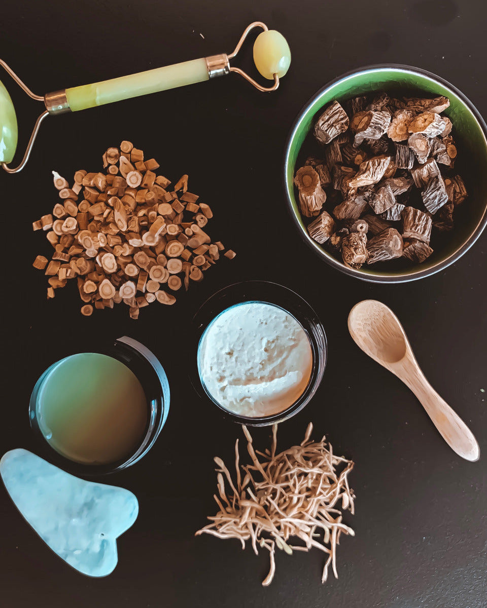 Collection of natural skincare products including a jade roller, bowl of dried herbs, and other items on a dark surface.